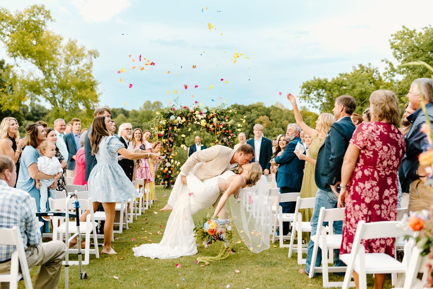 Outdoor wedding ceremony with guests seated on white chairs under blue sky with flower petals in air.