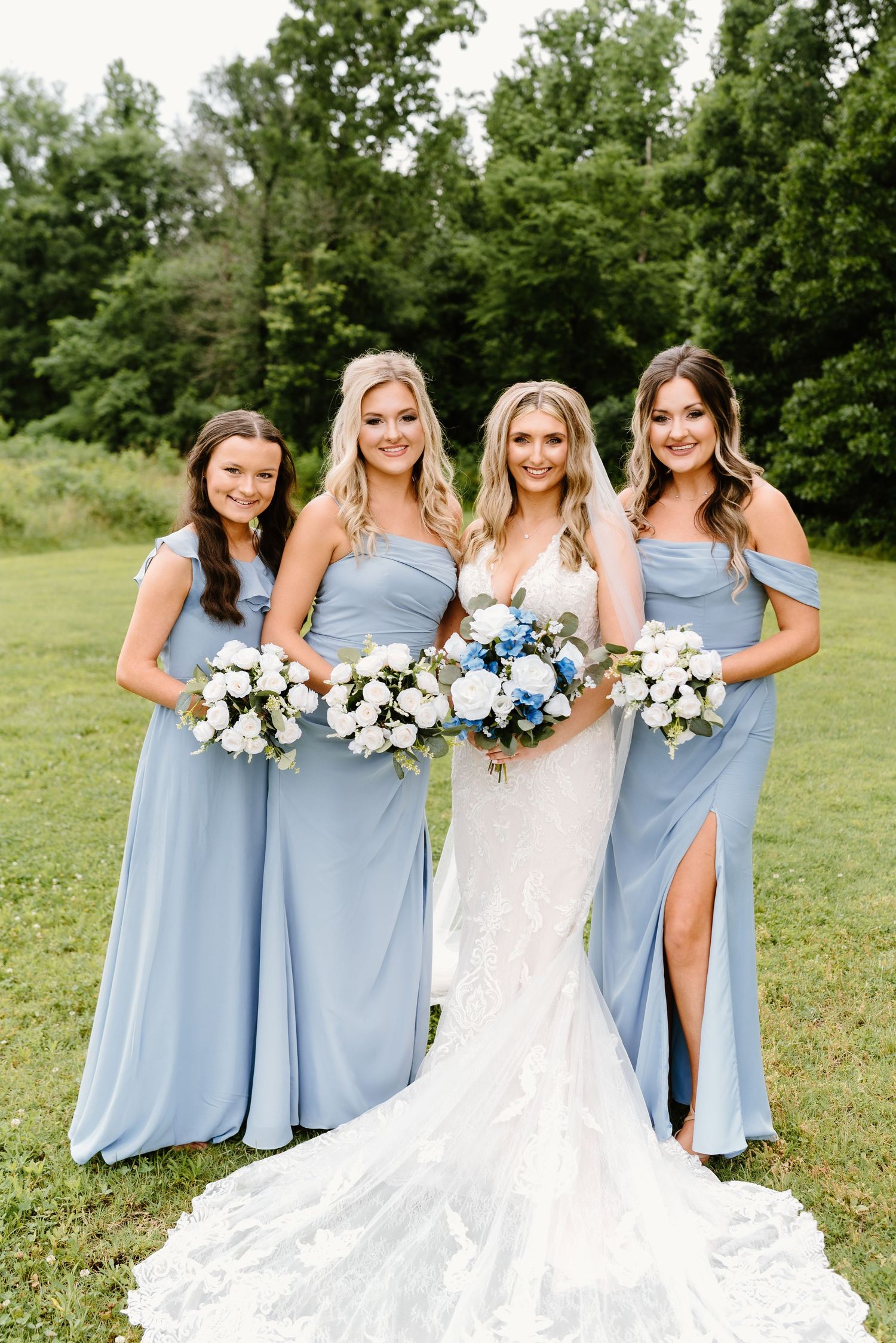 Bridesmaids in light blue dresses holding white bouquets pose together on a grassy lawn.