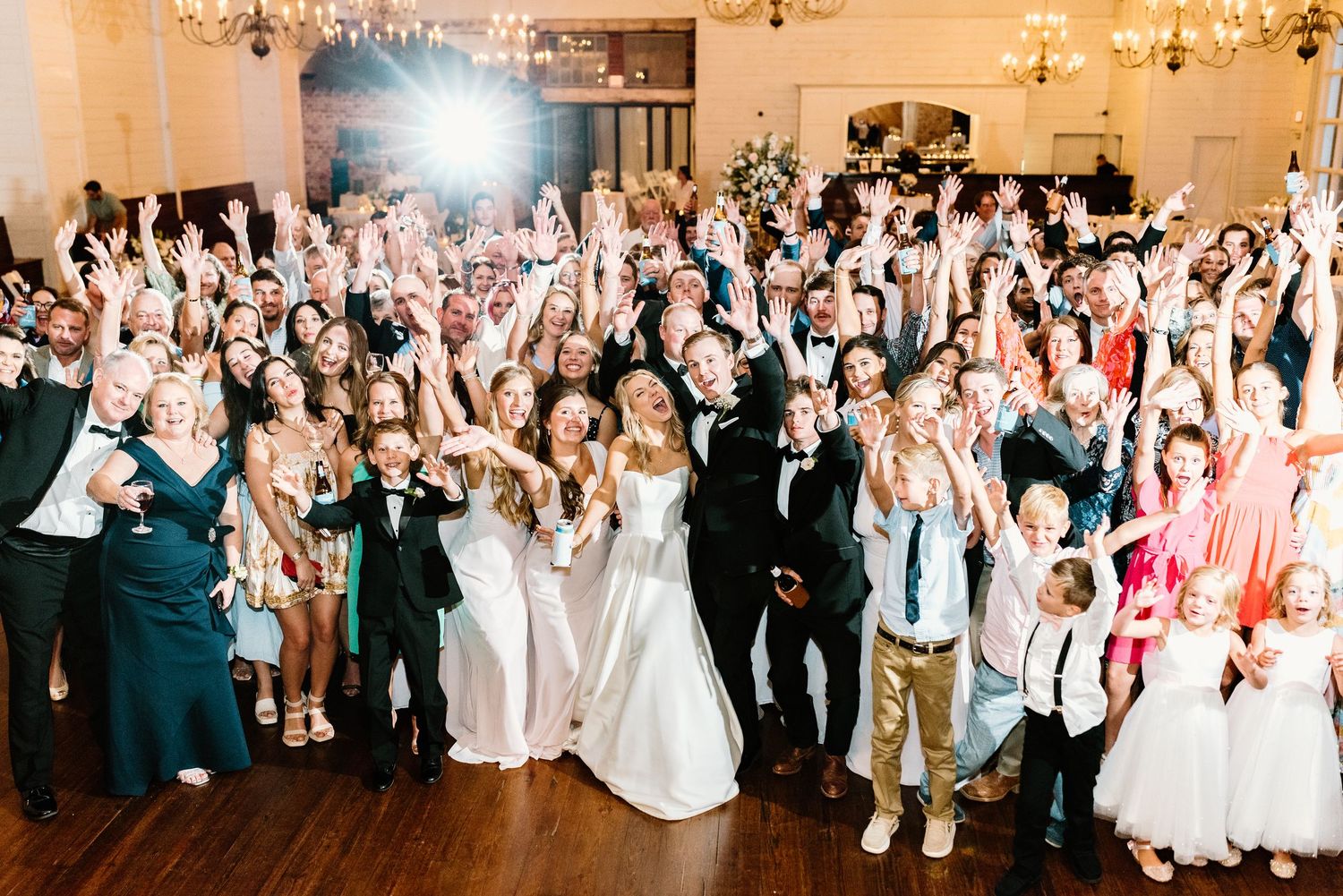 Wedding reception crowd dances and celebrates together in an elegant white ballroom with chandeliers.