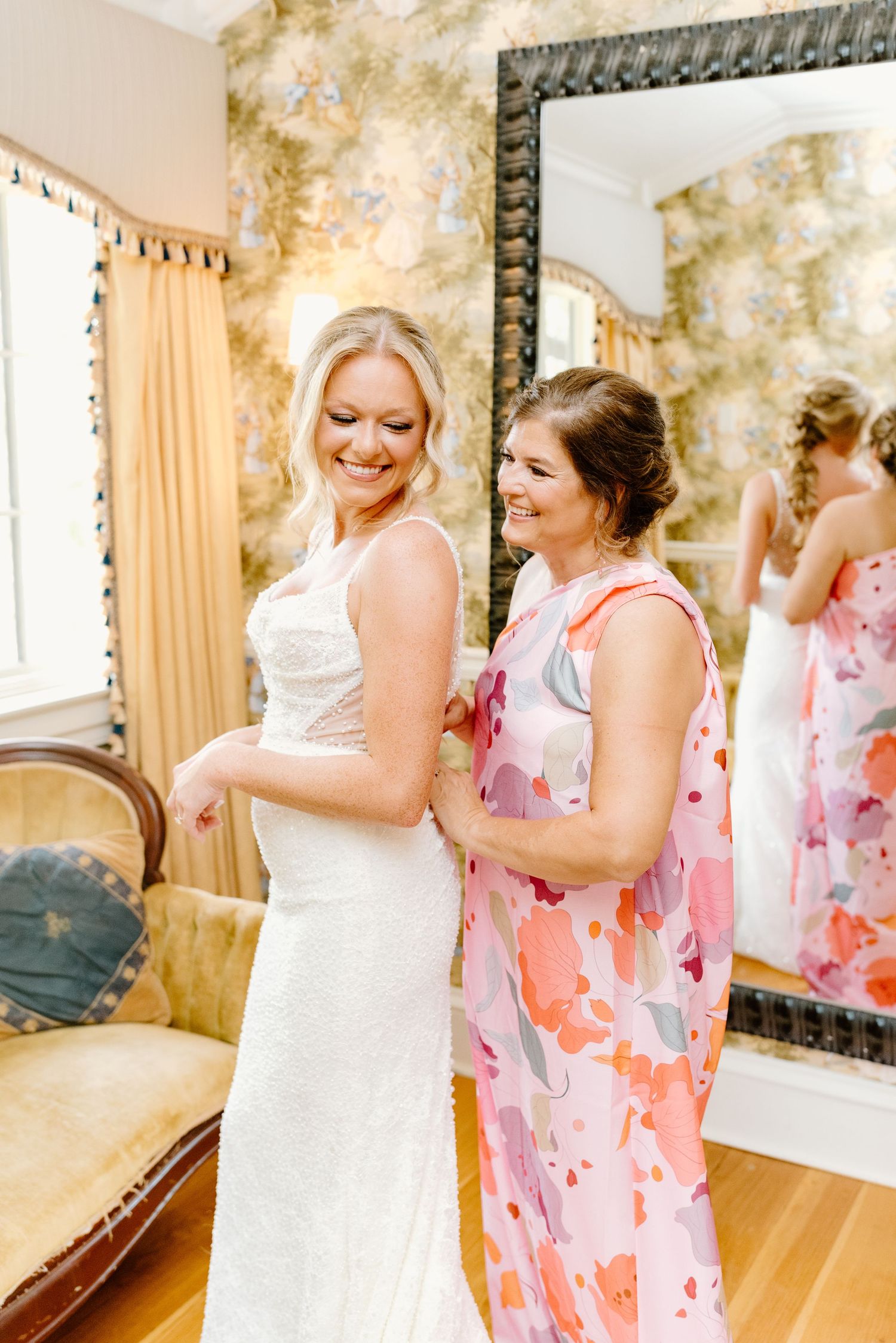 A bride in a white wedding dress shares joyful moments with attendants wearing pink floral dresses during wedding preparations.