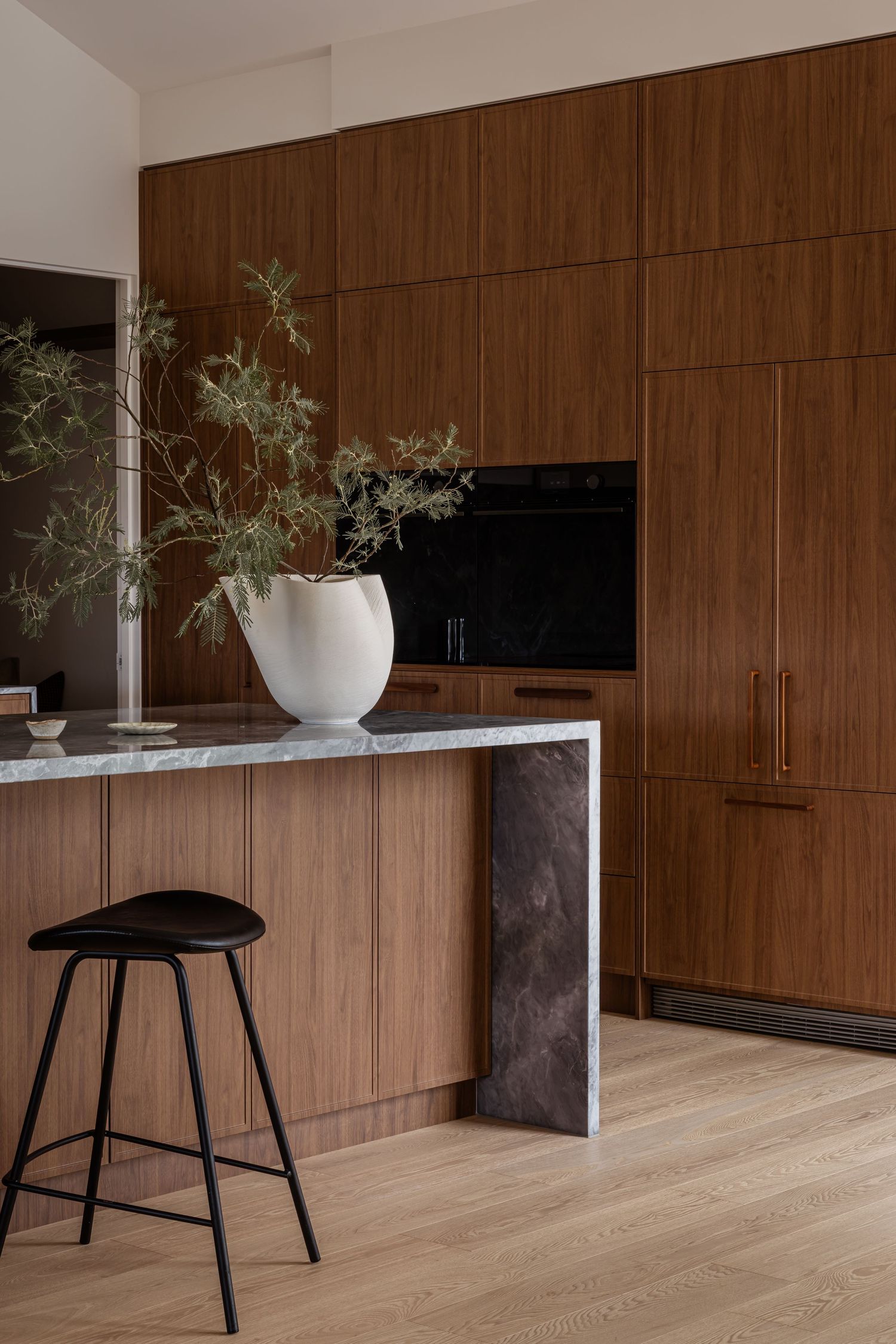 Modern kitchen interior with wood paneling, marble counter, and black bar stool beside potted plant.