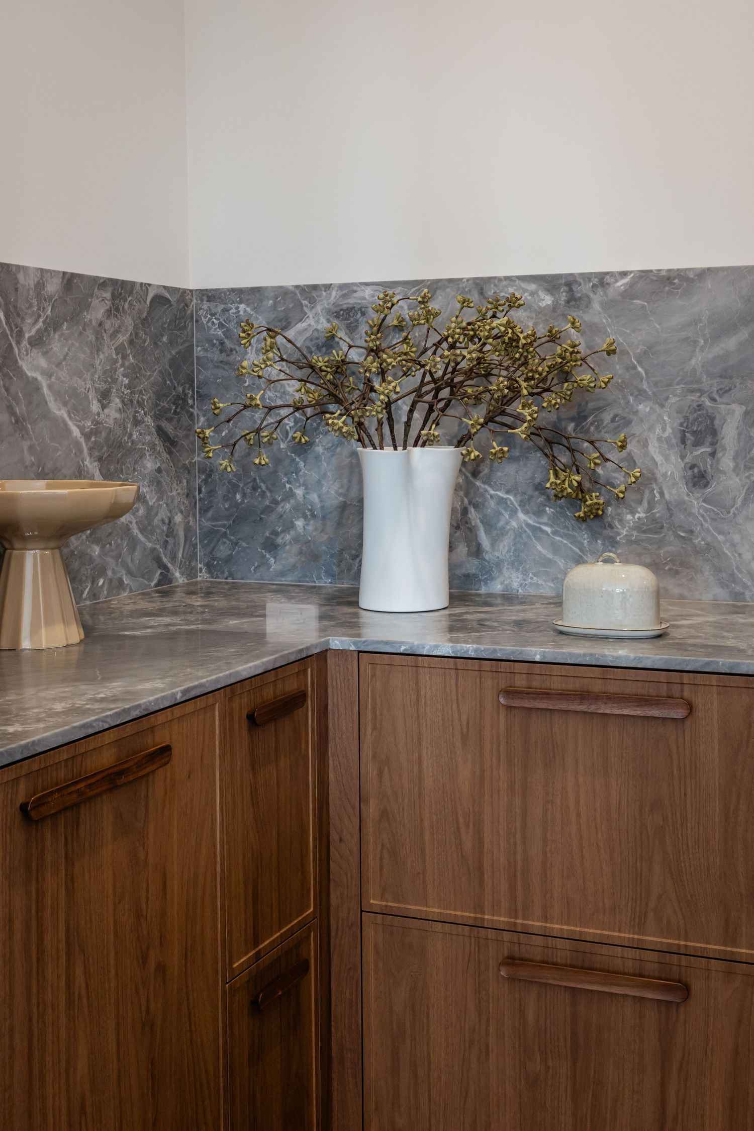Modern kitchen corner featuring walnut cabinets, marble countertops, and dried flowers in a white ceramic vase.