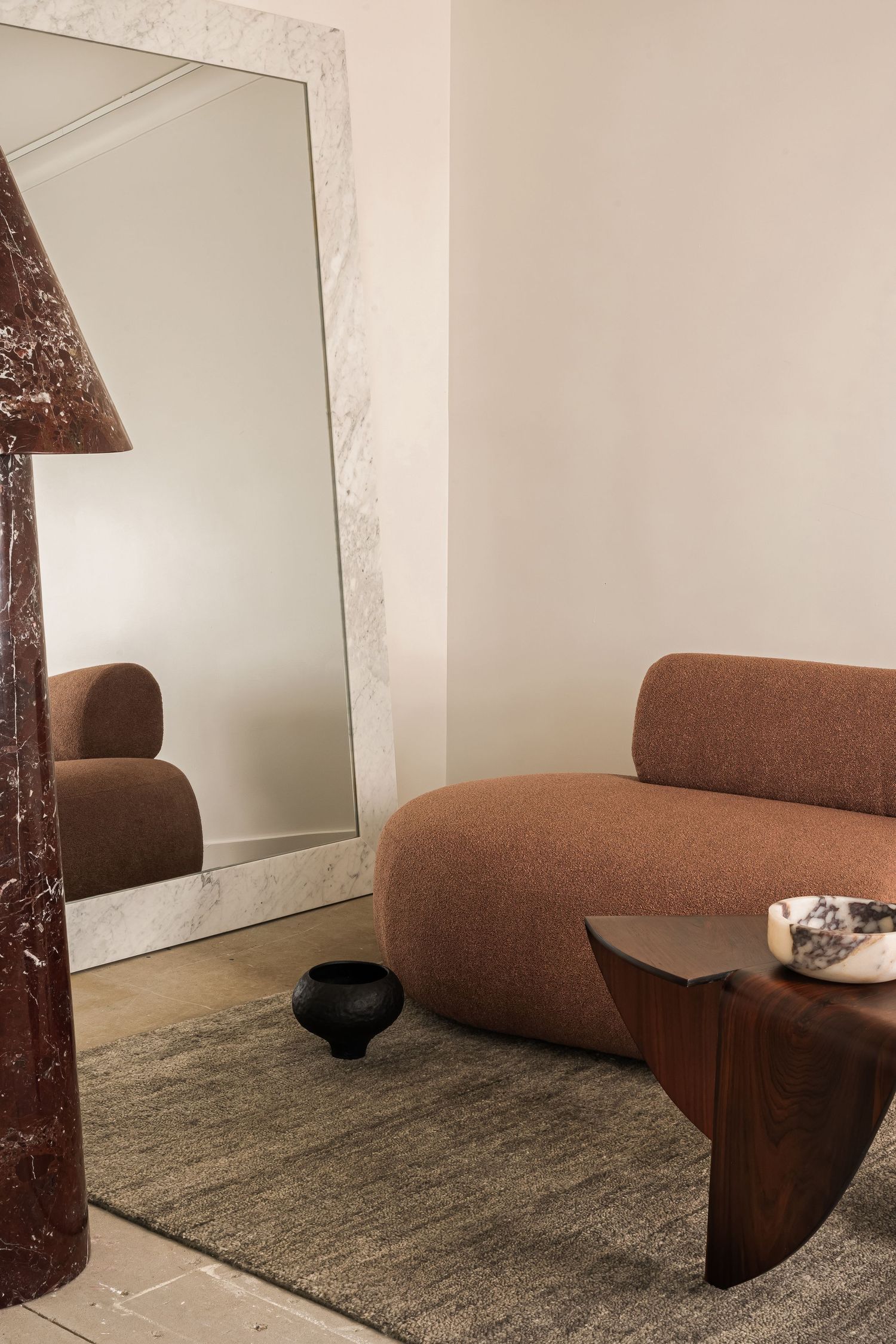 Contemporary brown curved chair positioned against white wall with decorative mirror on gray carpet.