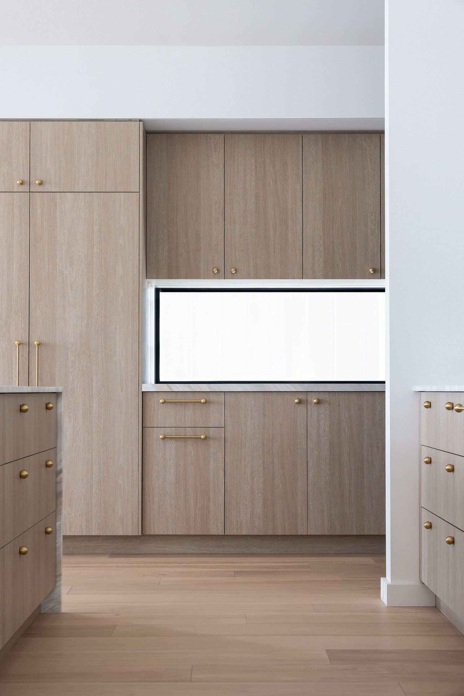 Modern kitchen interior with light wood cabinets, brass handles, and a horizontal window between upper and lower cabinets.