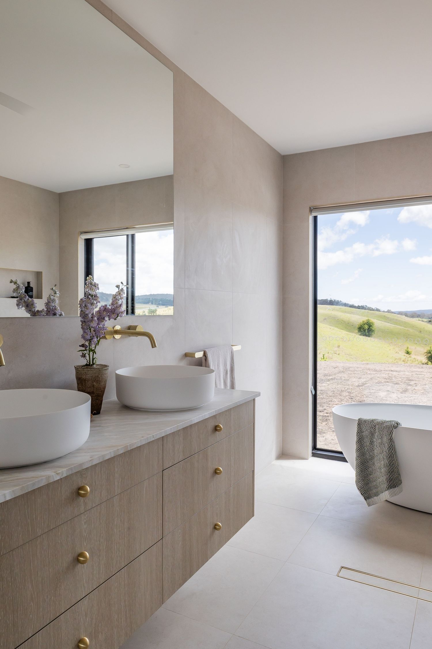 Modern bathroom with white vessel sinks, brass fixtures, and panoramic window overlooking rolling hills and countryside views.