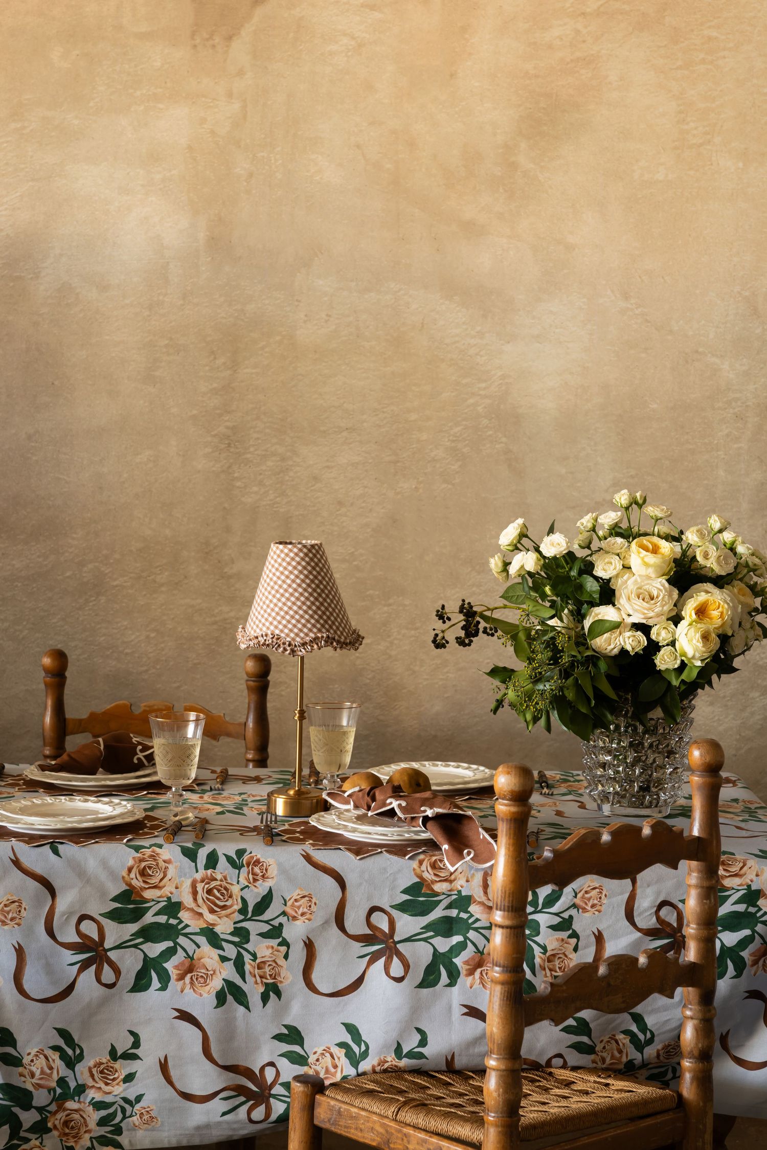 Vintage-style dining table setting with floral tablecloth, wooden chairs, and fresh yellow roses in crystal vase.