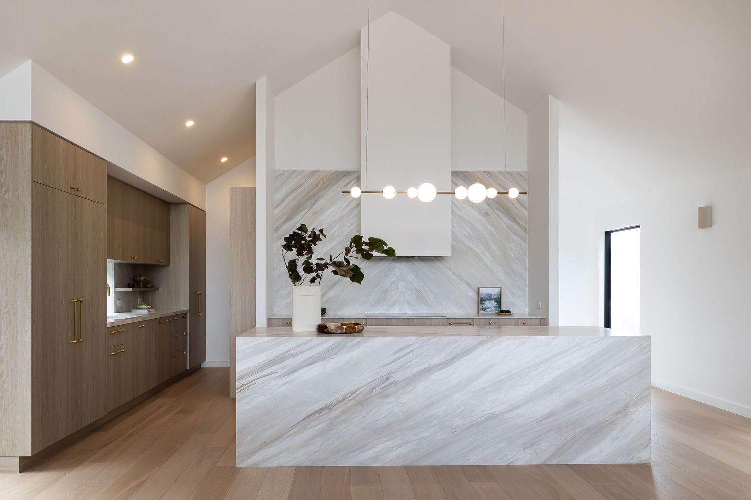 Modern kitchen design featuring white marble island and countertops with wood cabinetry and minimalist white range hood.
