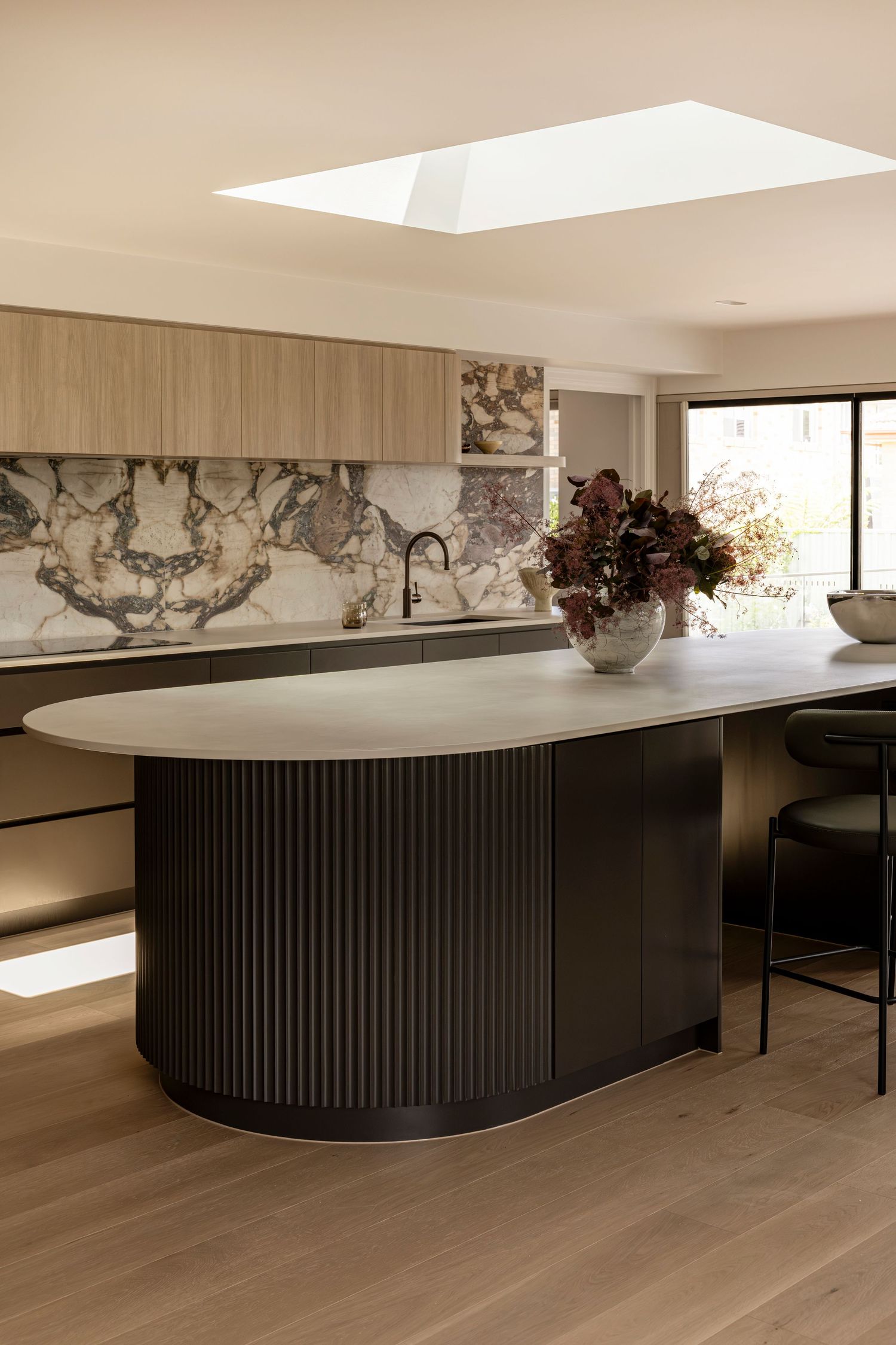 Modern kitchen design featuring a curved black island with ribbed panels and light wood flooring under skylight illumination.