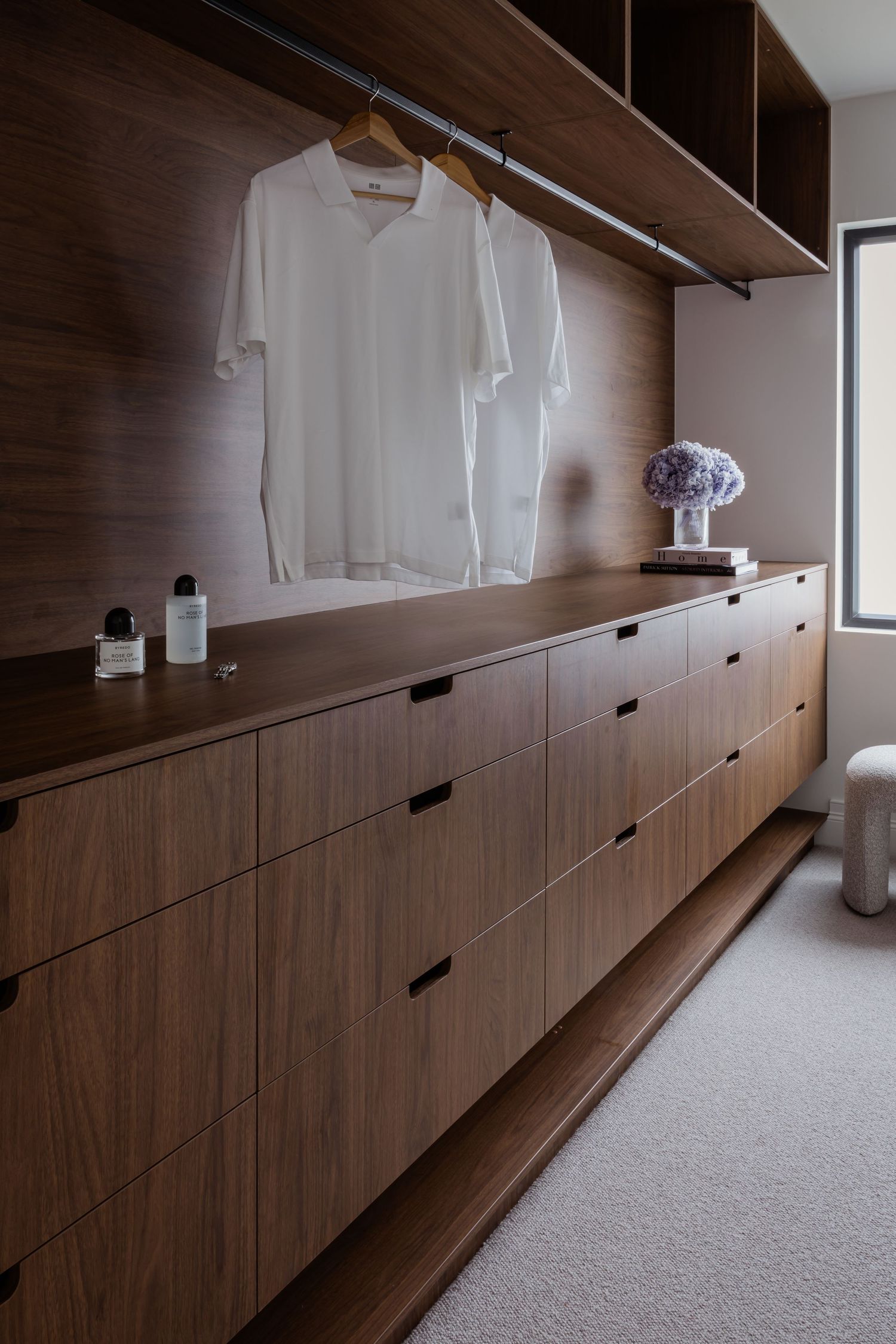 Long wooden wardrobe with drawers and hangers displaying a white shirt, with carpeted flooring and minimal decor.