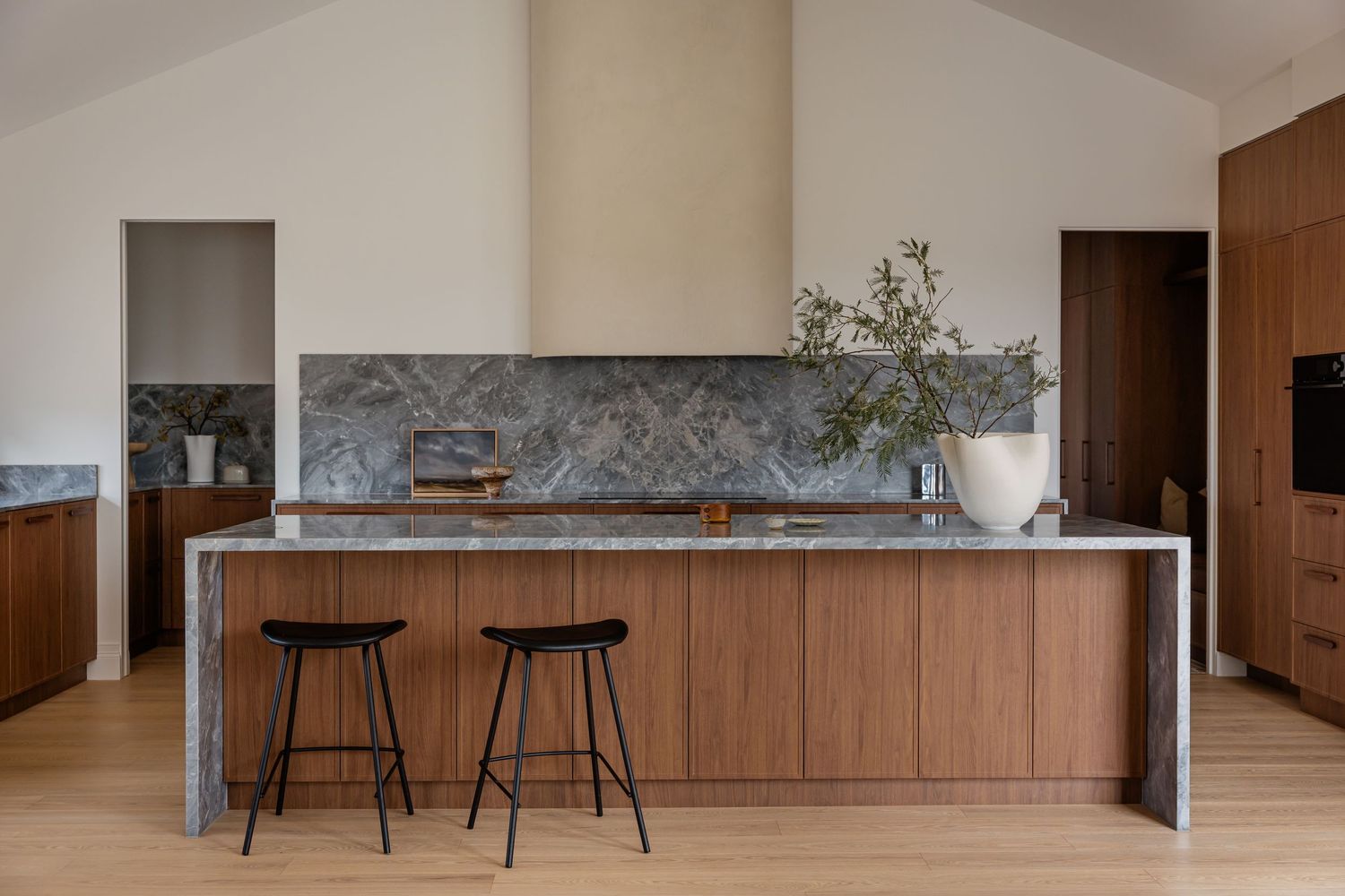 Modern minimalist kitchen with wood cabinetry, marble backsplash, waterfall island, and black bar stools.