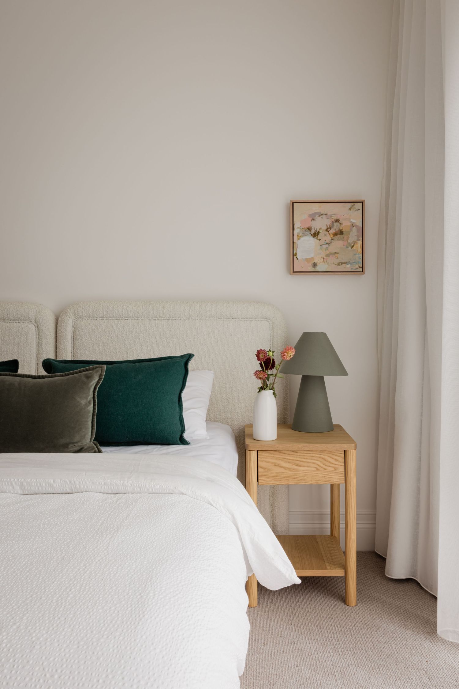 Minimalist bedroom with white bedding, emerald green accent pillows, wooden nightstand and grey table lamp on beige carpet.
