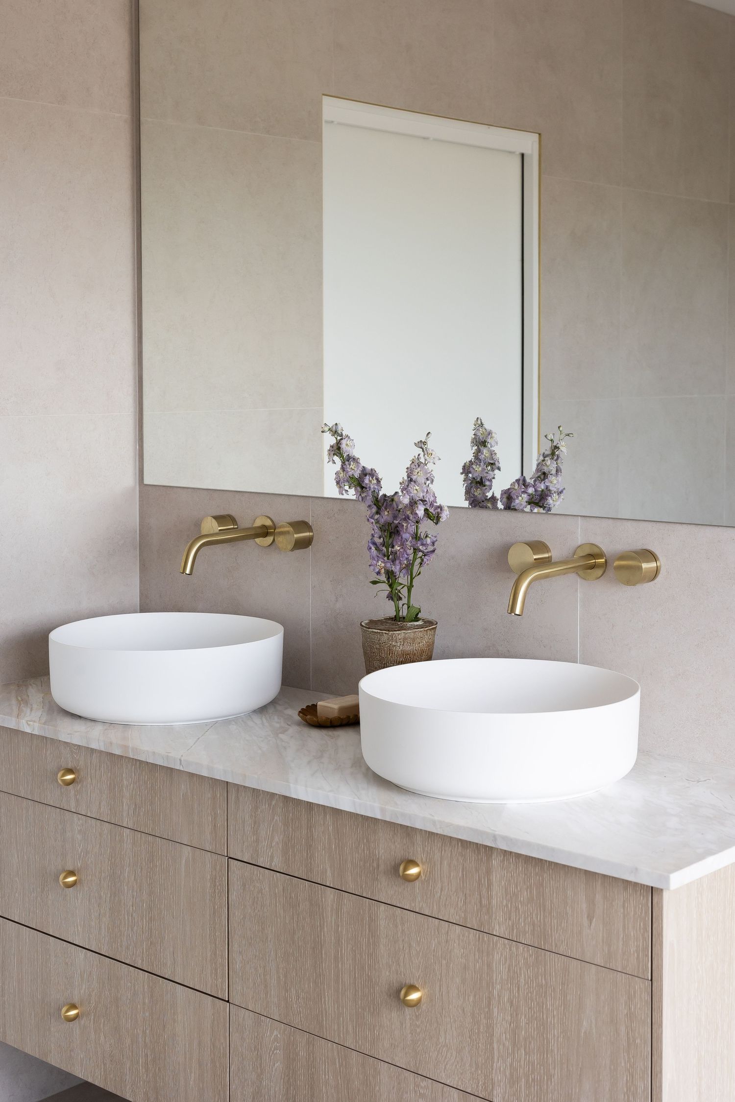 Modern bathroom vanity with white vessel sink, brass hardware, and light wood cabinetry against a textured gray wall.