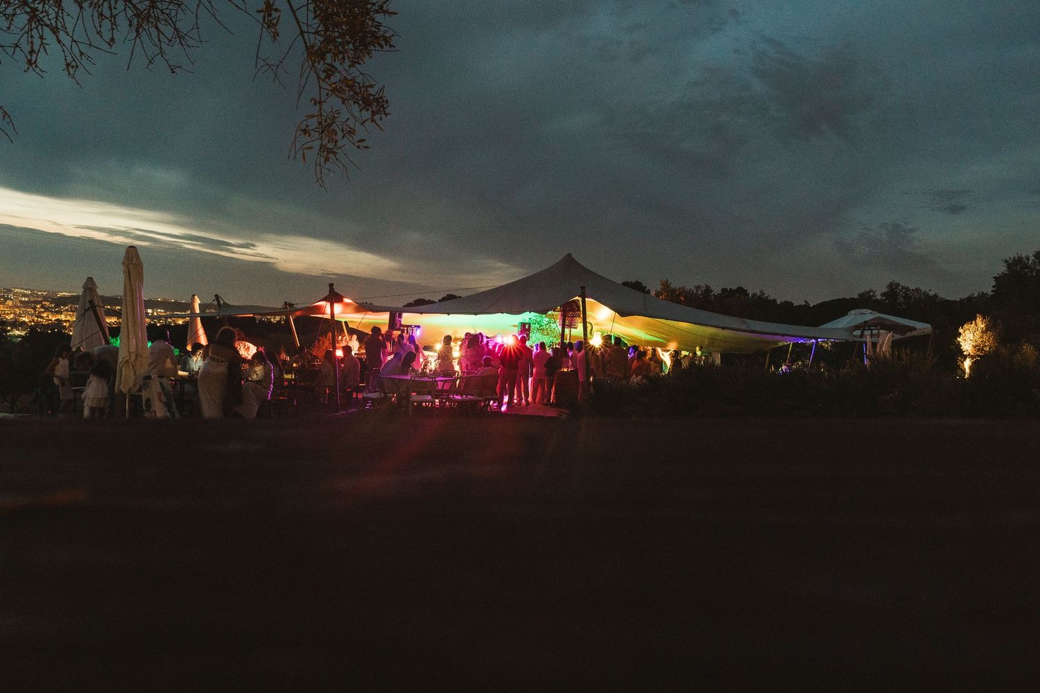Outdoor festival venue silhouetted against a dusky sky with colorful lights glowing in the distance.