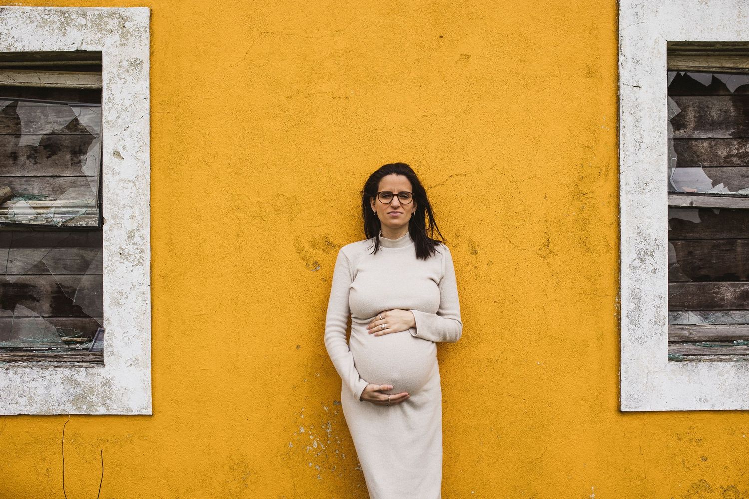 Maternity photos against a vibrant yellow wall with white window frames.