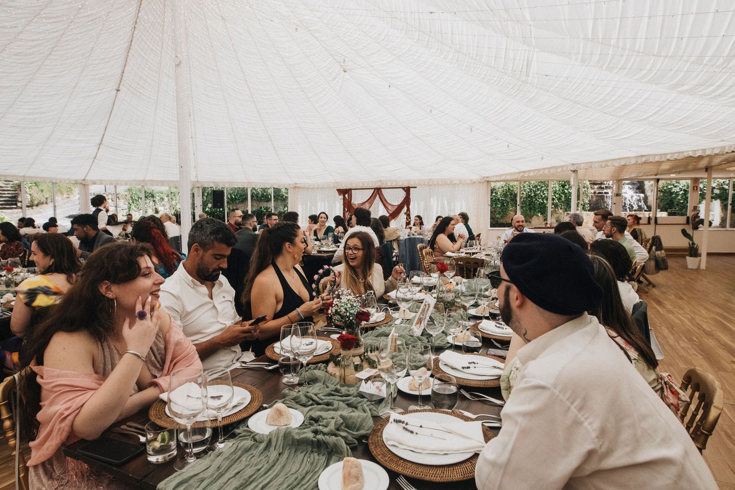Wedding reception tables decorated with greenery and place settings under a white tent.