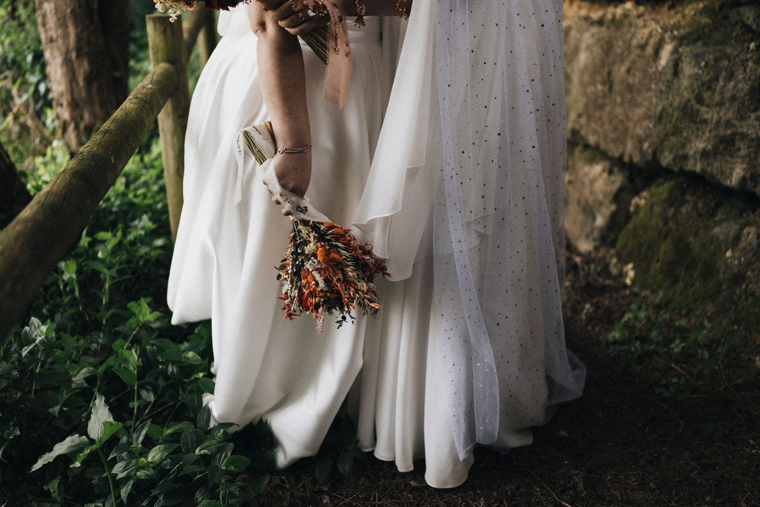 Row of white bridesmaid dresses hanging on rustic wooden hangers against stone wall.