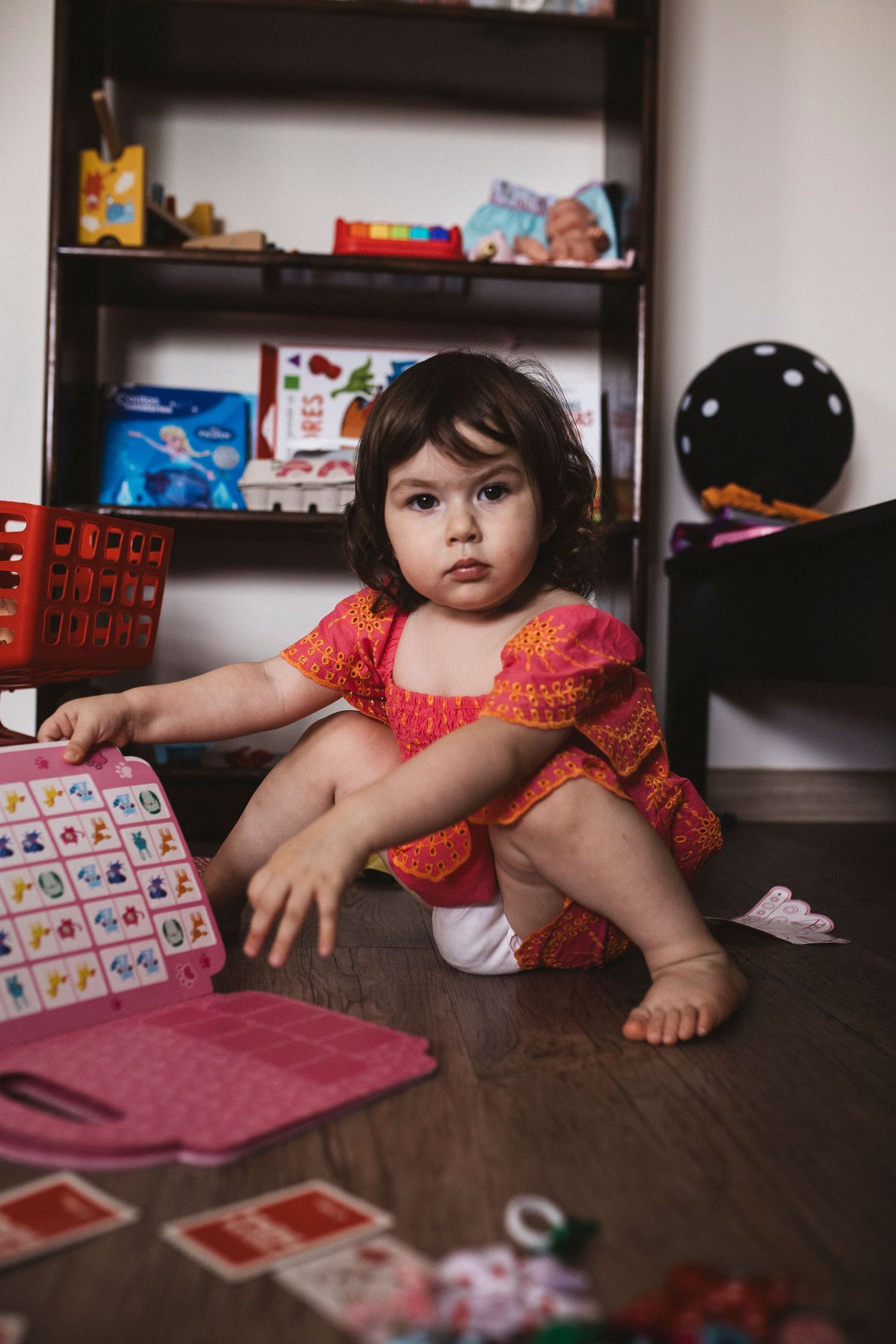Child in coral dress playing with cards on dark wooden floor near shelving unit.