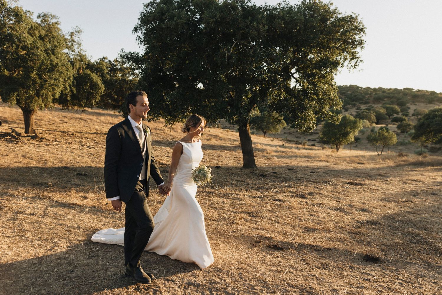 Newlyweds walk hand in hand through a golden field at sunset.