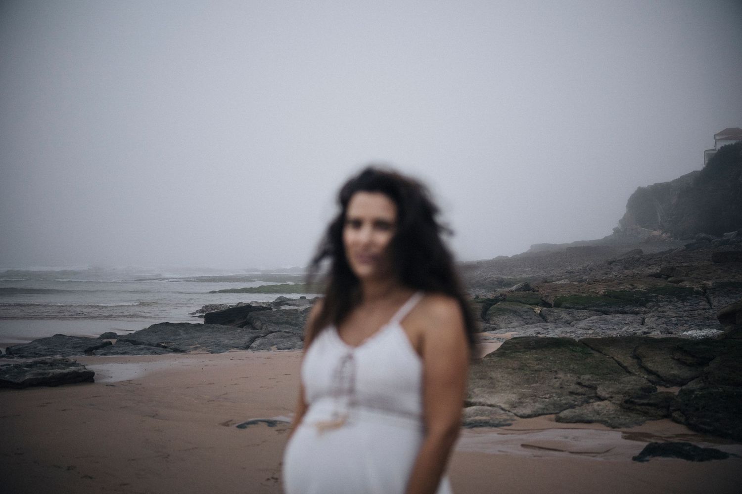 Atmospheric beach maternity photo shoot with misty coastal rocks in the background.