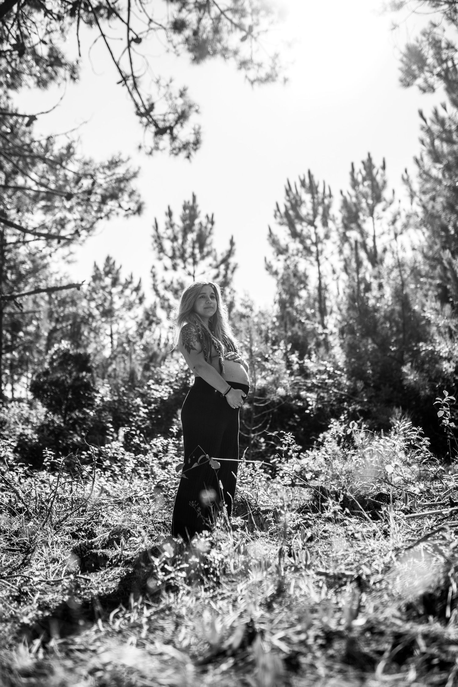 Artistic black and white maternity photo shoot in a sunlit forest with pine trees creating a dreamy atmosphere.