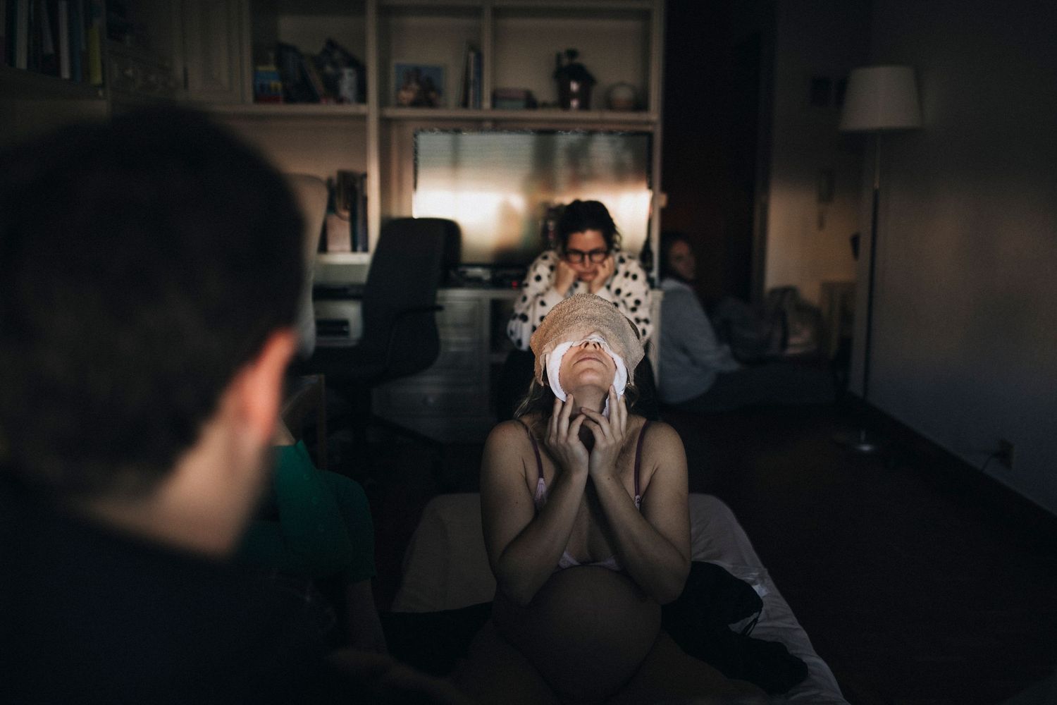 A person covers their face with hands while another person watches in a dimly lit living room.