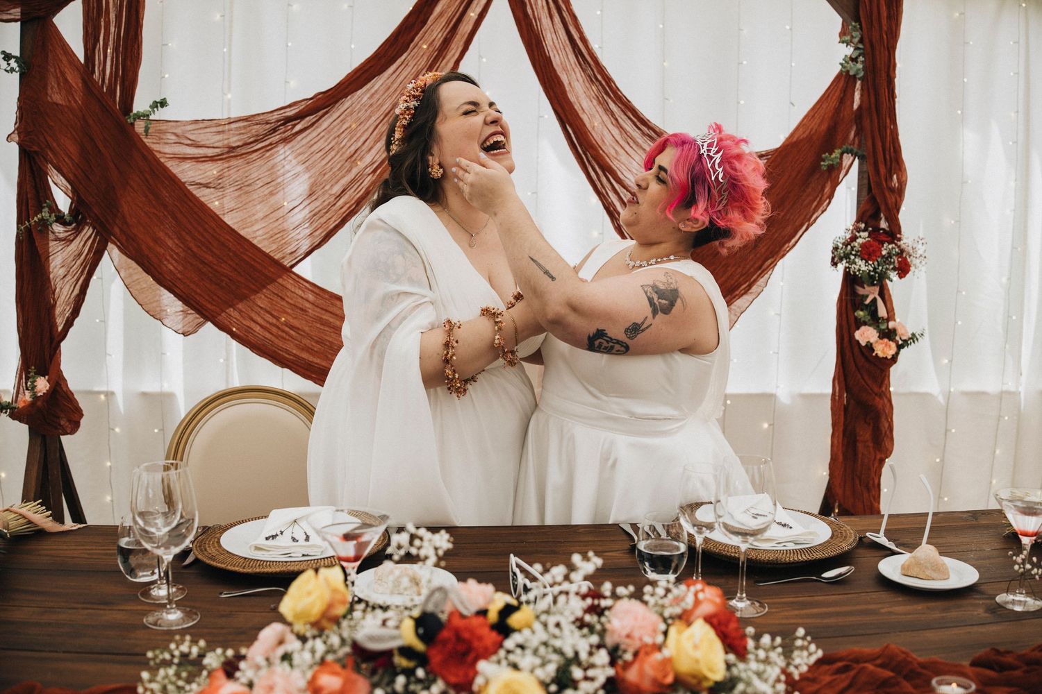 Two people in white dresses share a candid laughing moment at a decorated wedding reception table.