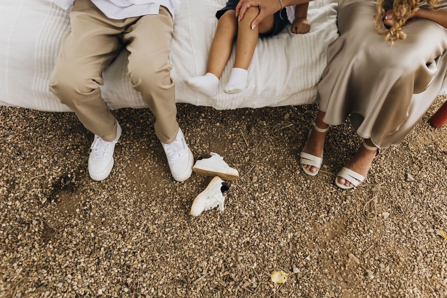 Three pairs of legs in casual attire sitting together on ground wearing white sneakers and sandals.