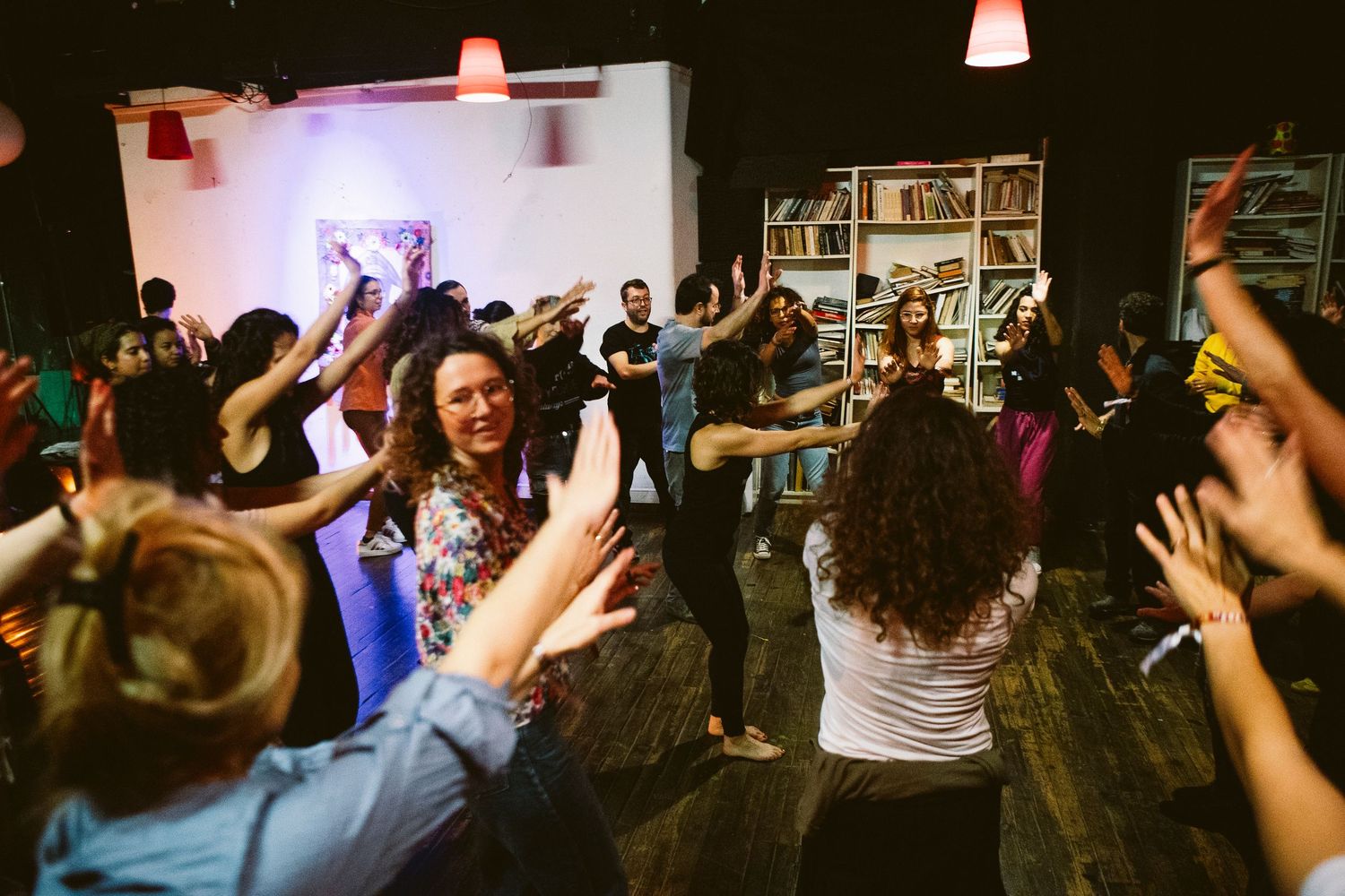 Crowd of people dancing with raised hands at a nightclub or party venue with red lighting overhead.
