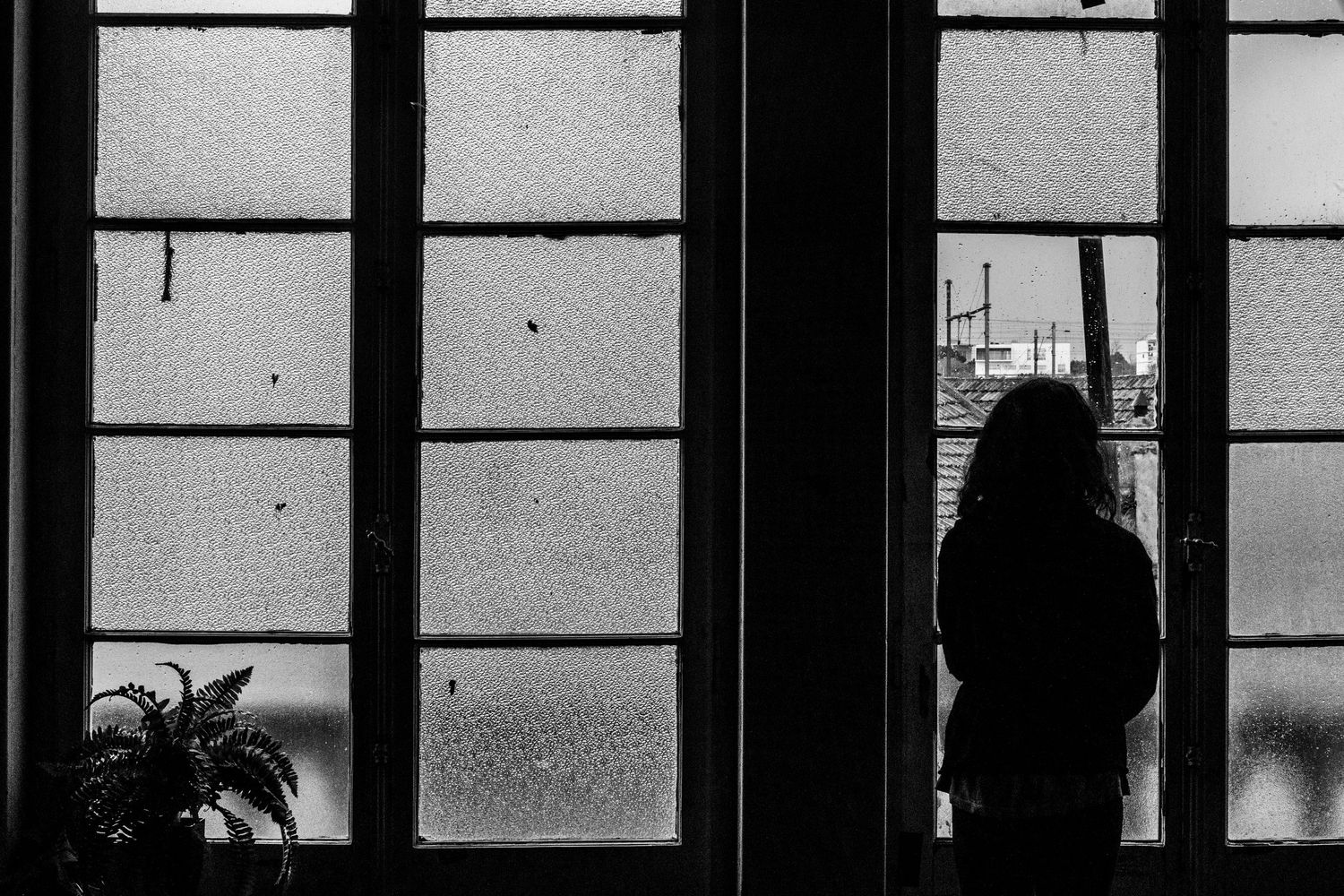 A silhouetted figure stands by a window with gridded panes in a moody black and white composition.