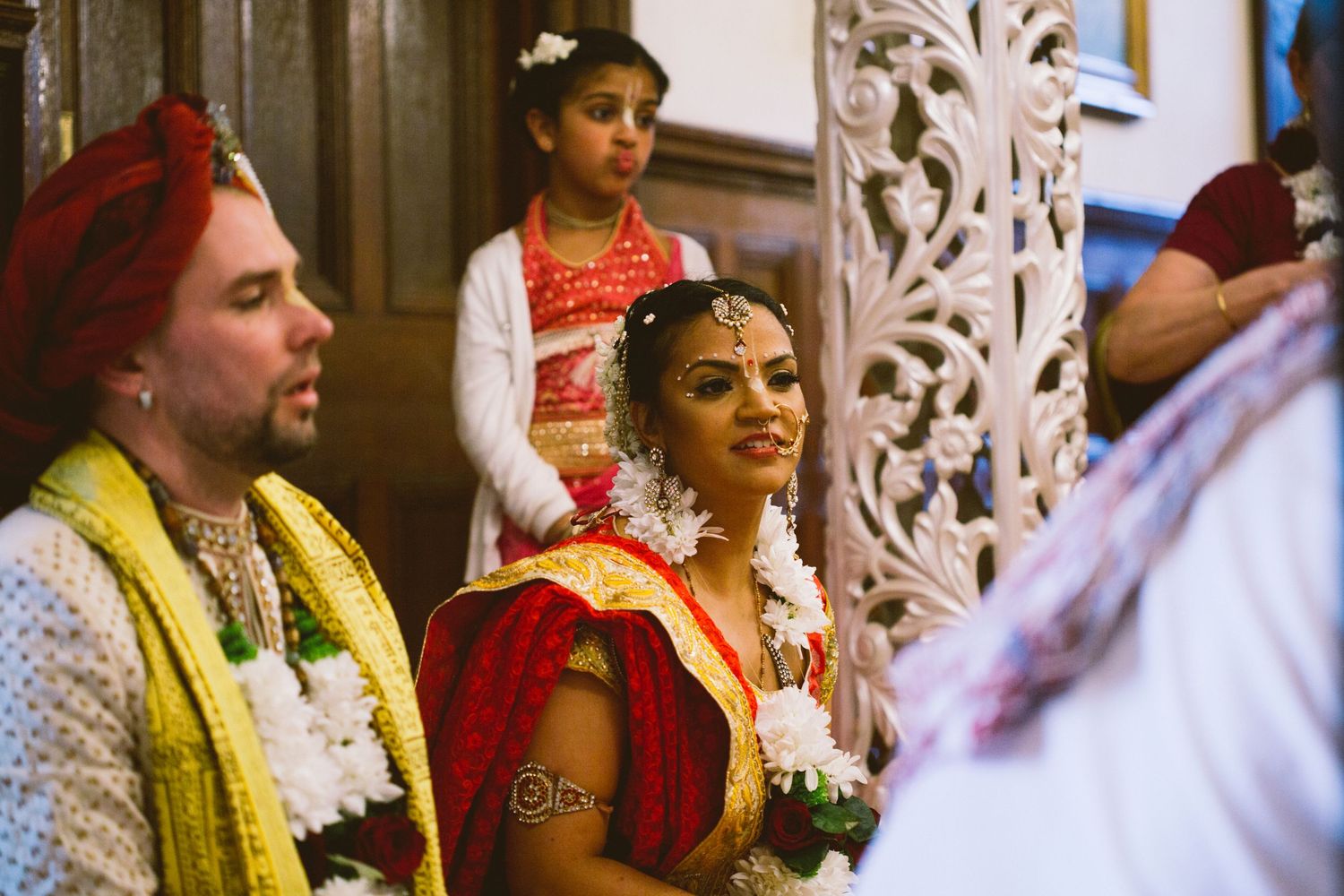 Couple in traditional Indian wedding attire seated during ceremony with ornate white backdrop and decorative elements.