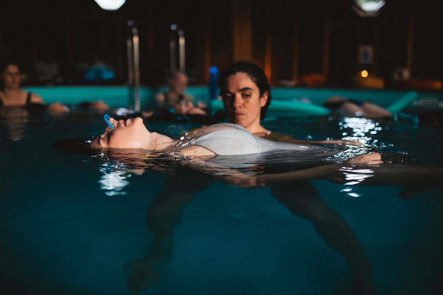 Swimmers in illuminated pool at night with turquoise water reflections.
