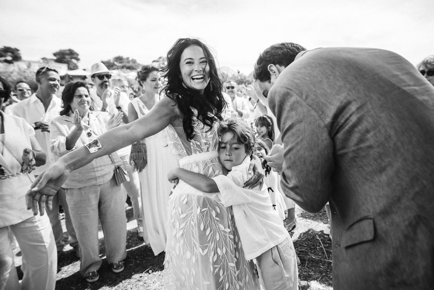 Joyful wedding guests celebrate outdoors in white attire with raised arms and excited expressions.