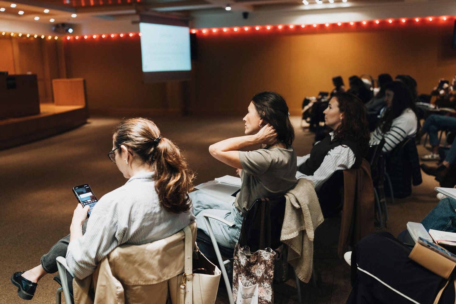 Students sit in rows at desks in a dimly lit lecture hall with presentation screens visible on the walls.