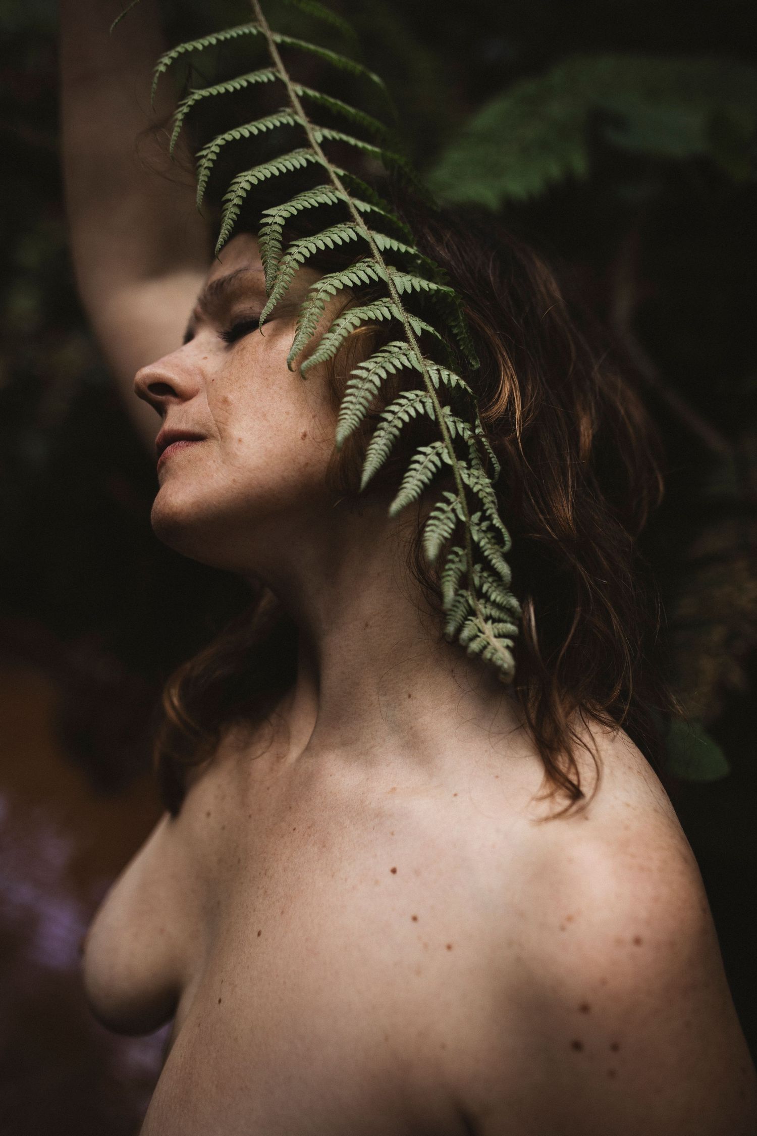 Delicate fern frond draped over bare shoulder in moody forest lighting.