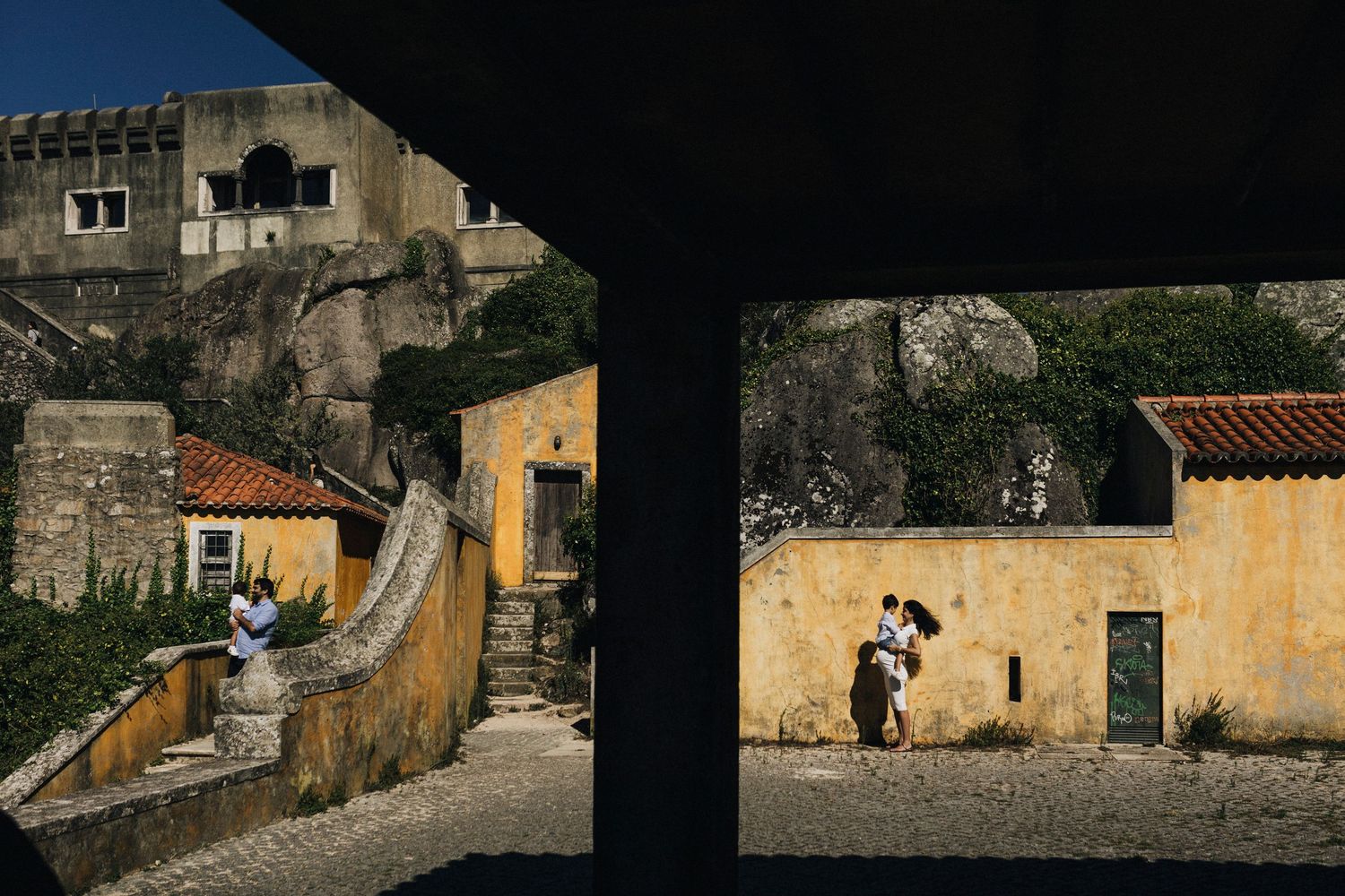 A couple poses beneath an archway in a rustic European village with yellow walls and stone architecture.