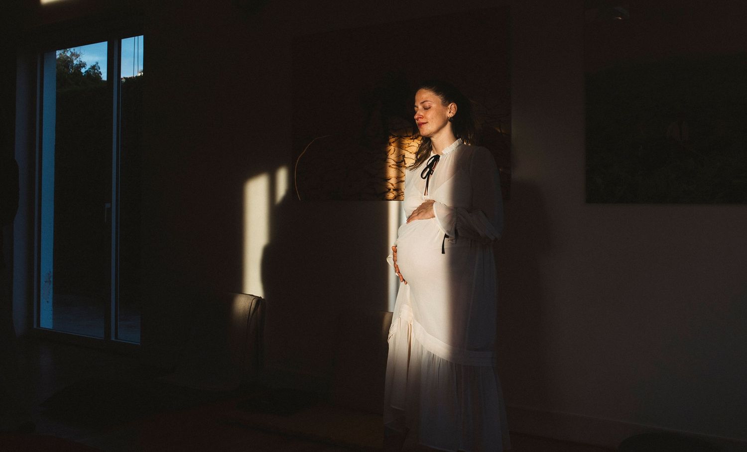 Dramatic black and white portrait showing a figure illuminated by window light in a dark interior space.
