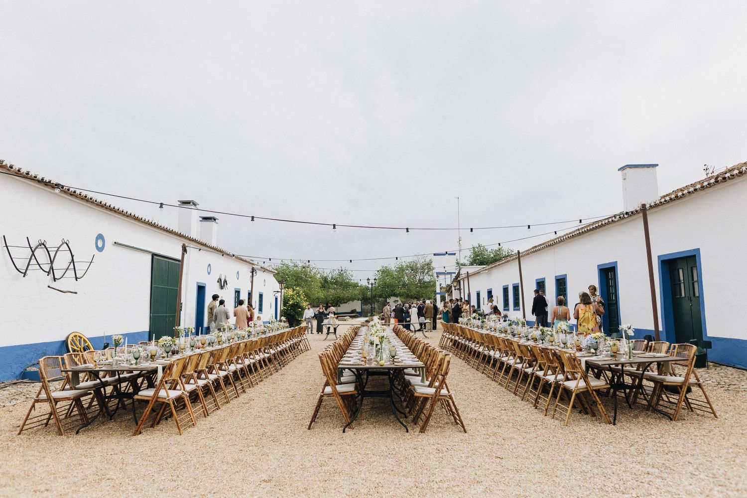 Long rustic outdoor dining tables set up between white buildings with blue trim and string lights overhead.