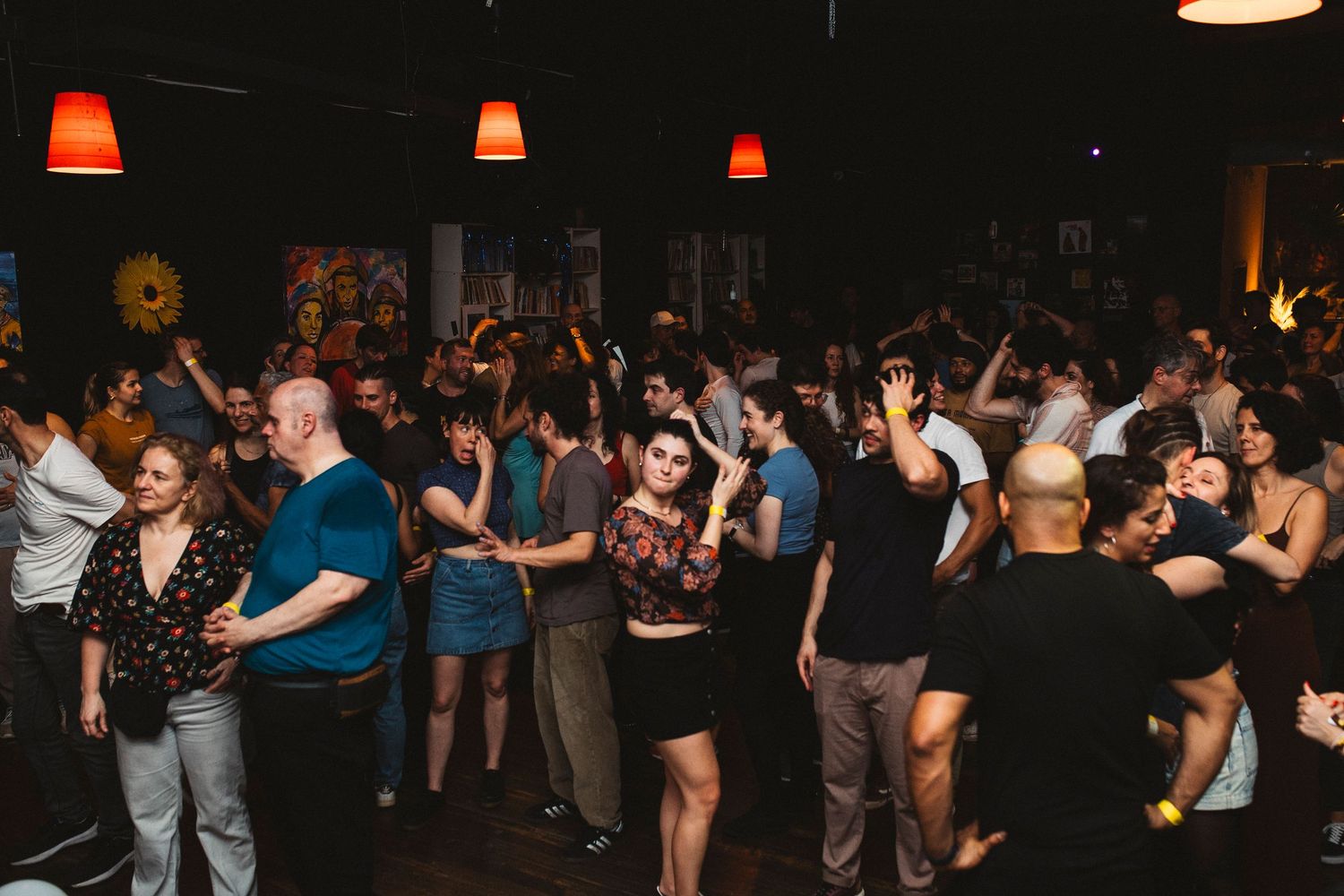 People dancing and socializing at a dimly lit nightclub with red pendant lights hanging from the ceiling.