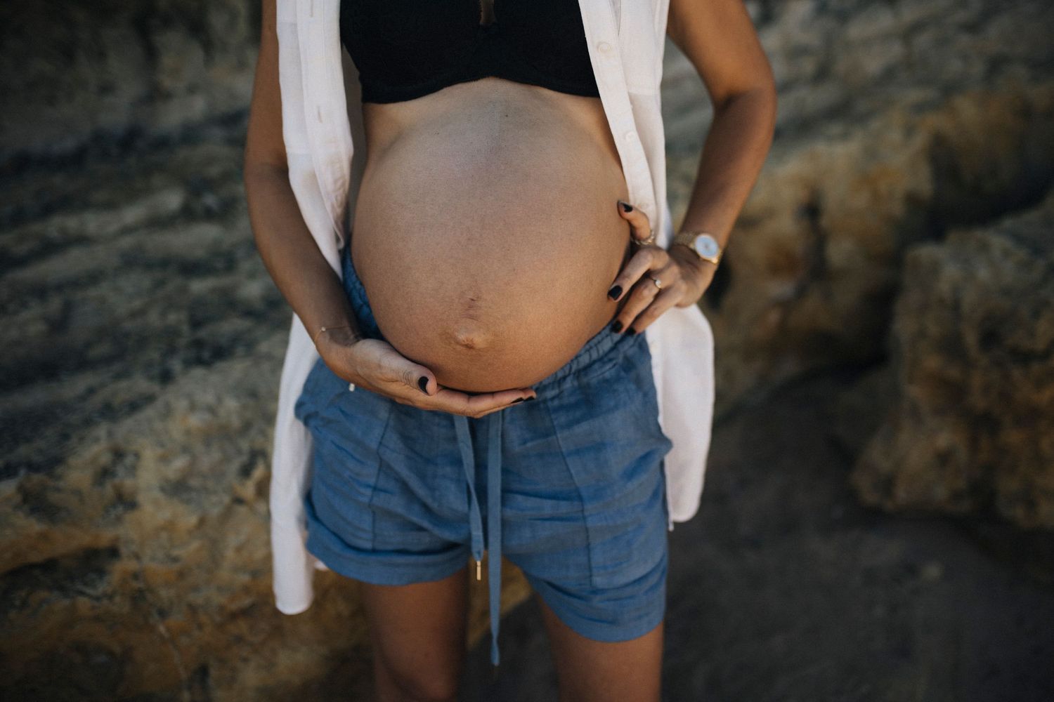 A pregnant person in blue denim shorts and white top poses against a textured rock wall background.