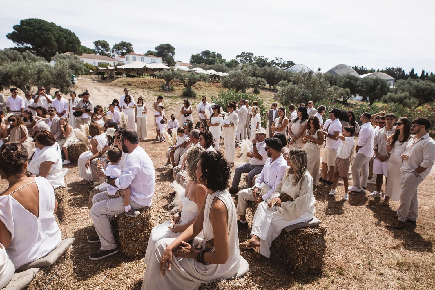 Wedding guests in white attire gather outdoors for a rustic celebration in a Mediterranean countryside setting.