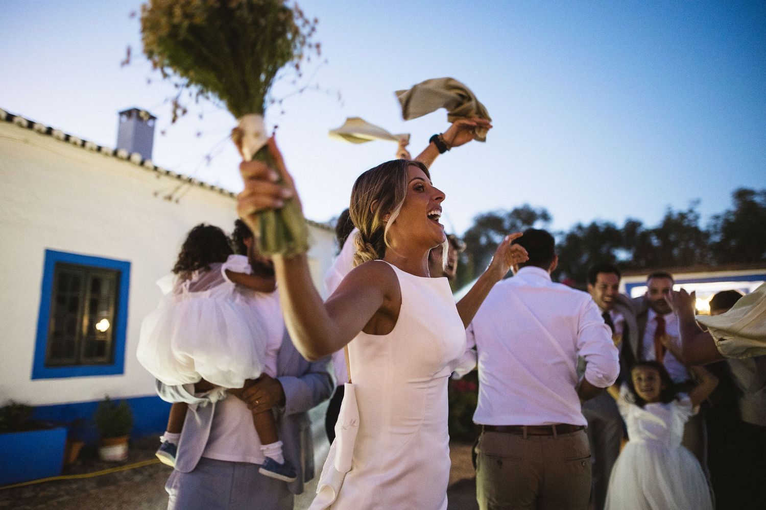 Wedding guest raises a bouquet in celebration during an outdoor reception at a villa with blue trim.