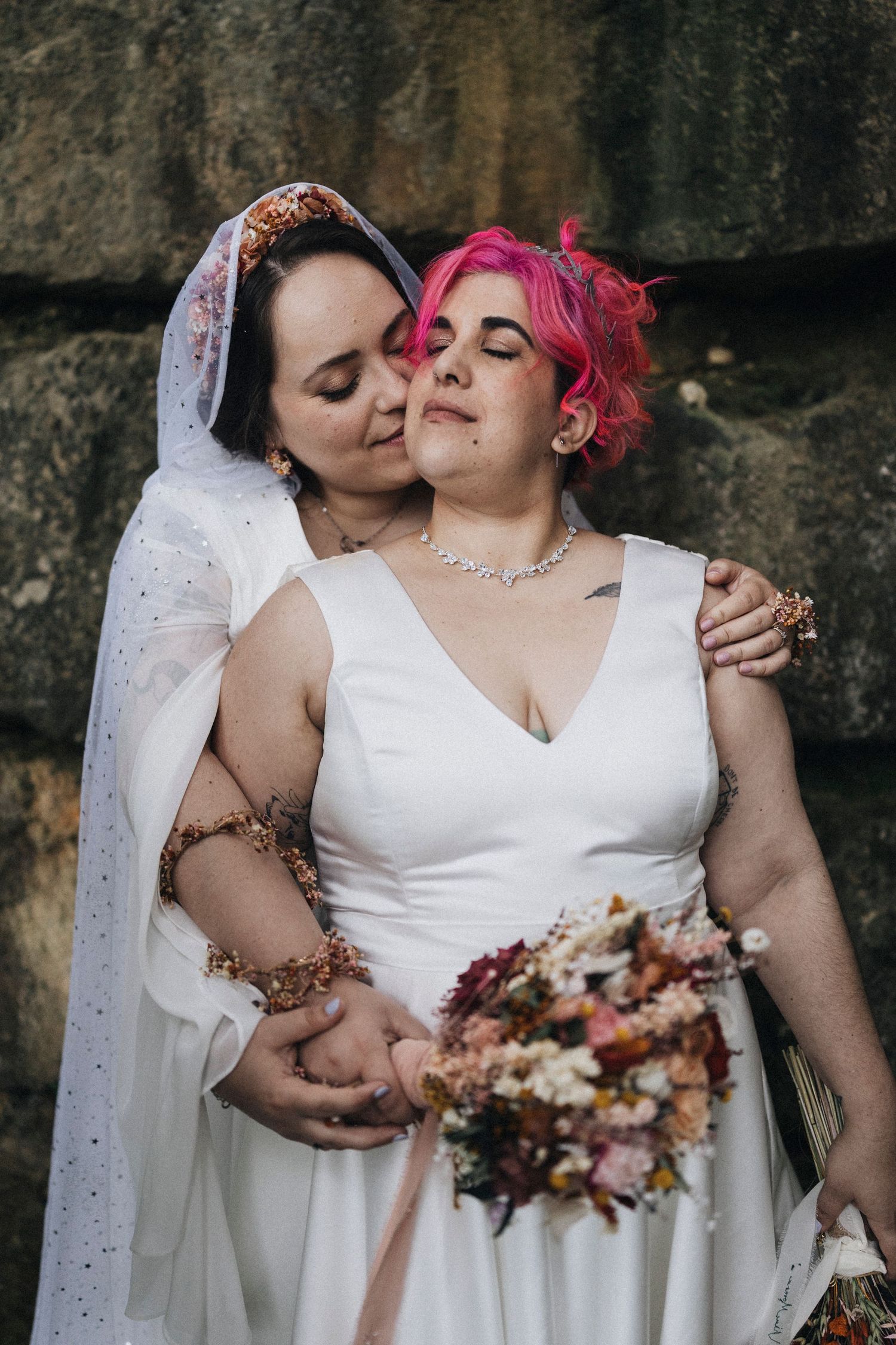 Two brides in white wedding dresses share an intimate moment against a stone wall while holding autumn-colored bouquets.