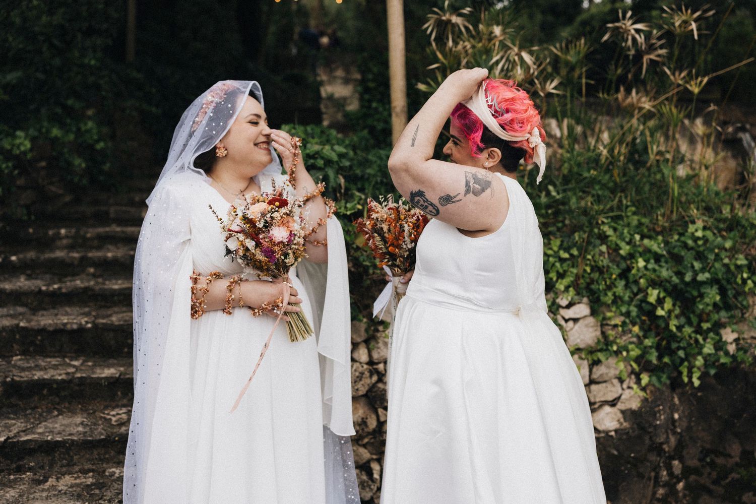 Two people in white wedding dresses share a joyful moment together in an outdoor garden setting.