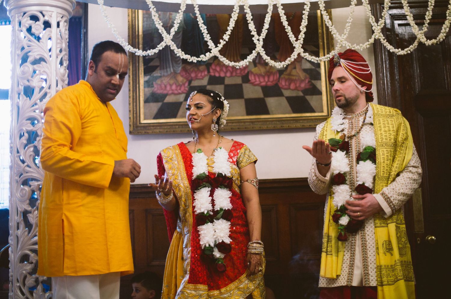 Three people in traditional Indian wedding attire stand together during a ceremony with decorative lights in background.