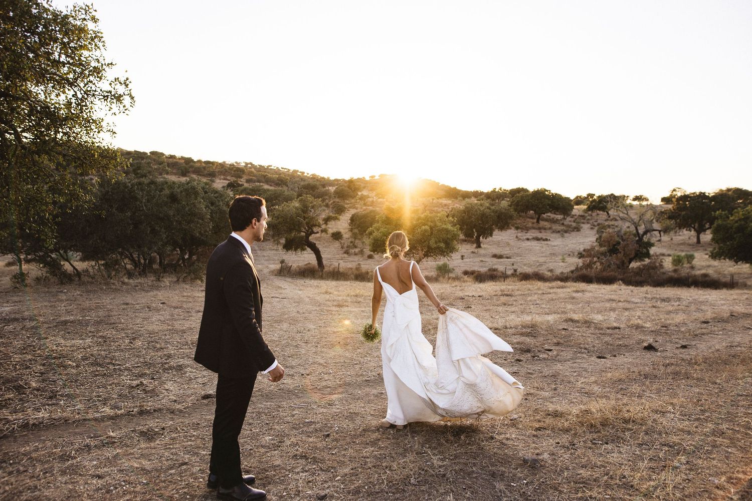 A couple in wedding attire stands in a desert landscape at sunset with the bride's dress flowing in the breeze.