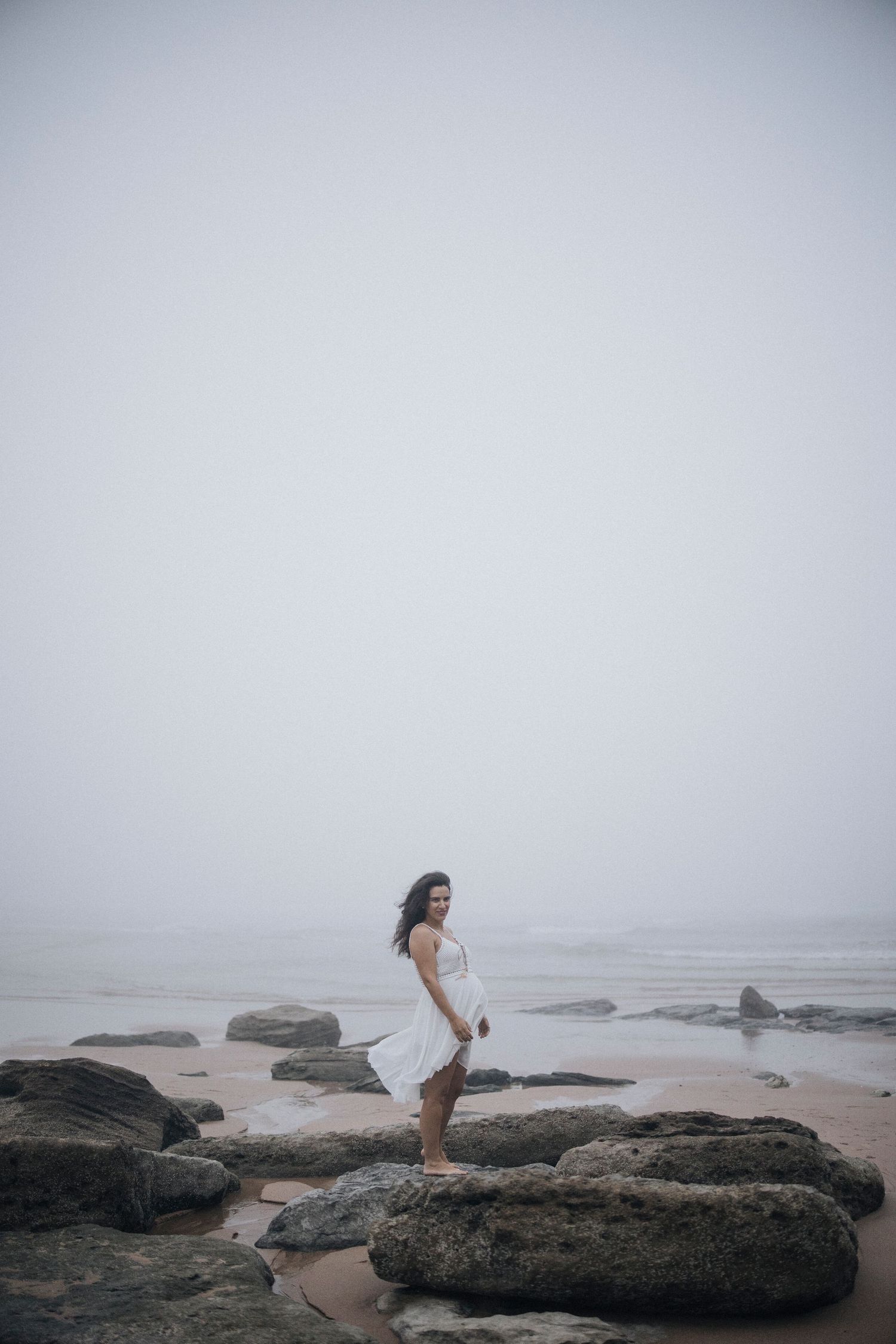 Dramatic coastal maternity photography with rocky shoreline and misty ocean backdrop.