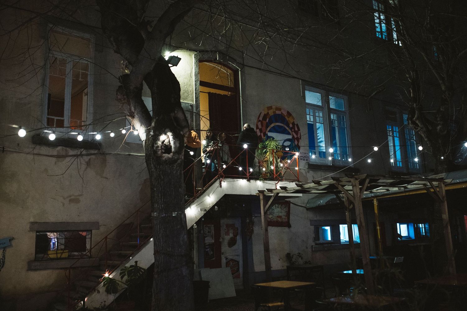 A dark nighttime scene shows a row of illuminated windows and string lights creating an atmospheric urban setting.