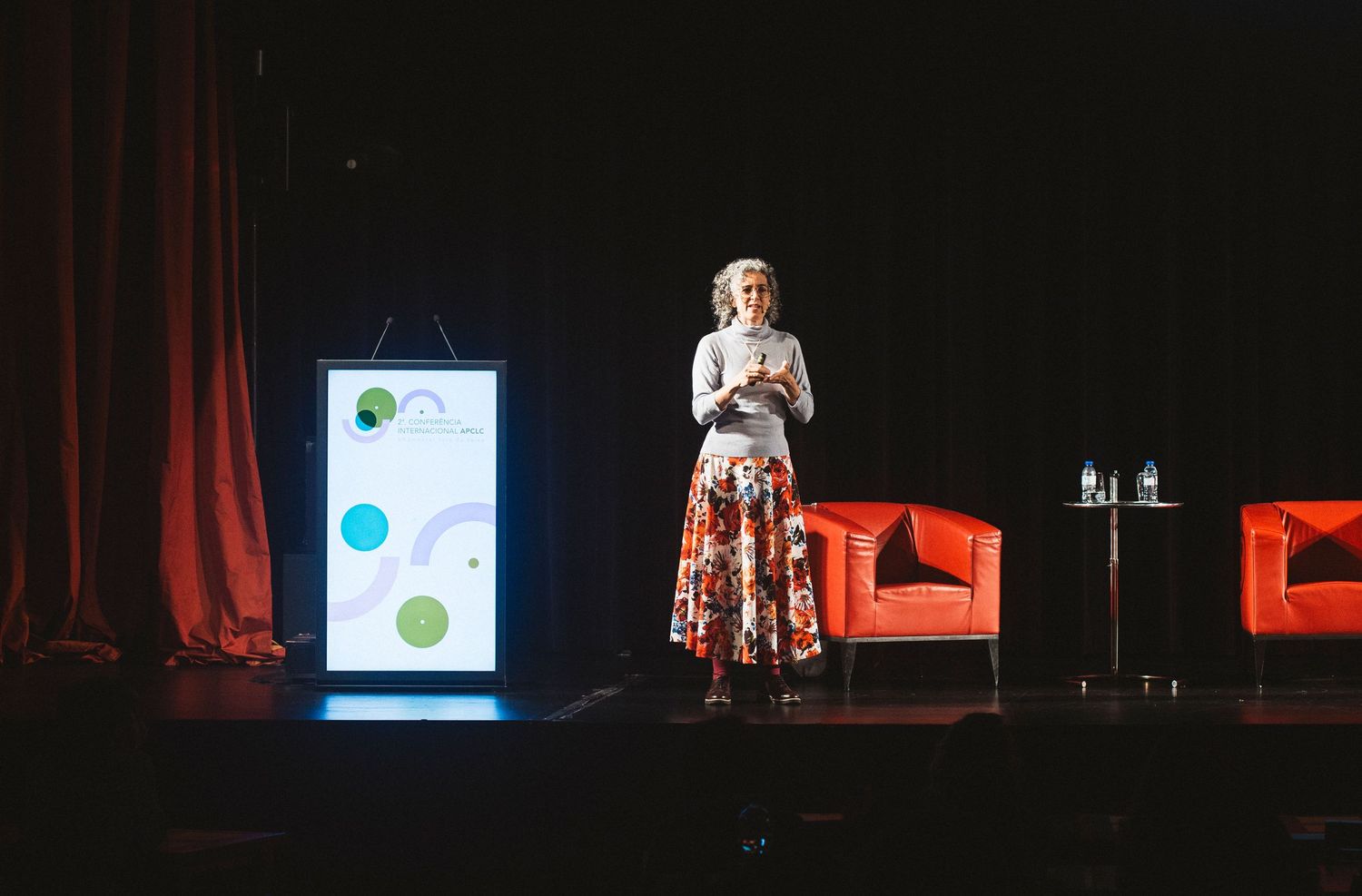 A dimly lit stage with red curtains and a presentation screen showing data visualizations in a dark theater setting.