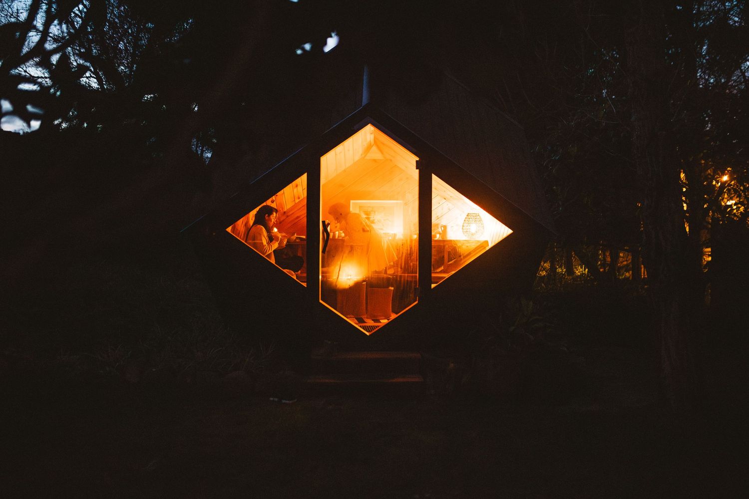 Orange diamond-shaped windows glow at dusk through trees creating a geometric pattern.