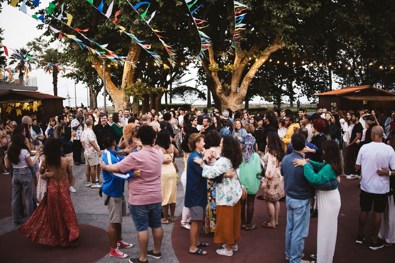 A crowd of people gather outdoors under trees during a social event at dusk.